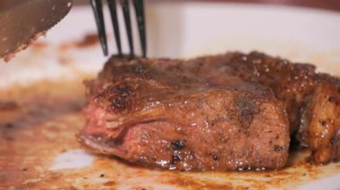 Tabakta büyük bir parça taze ızgara biftek. Ağır çekimde kanla birlikte orta pişmiş et. Bir çatal ve bıçak yakın görüntü kesiyor.