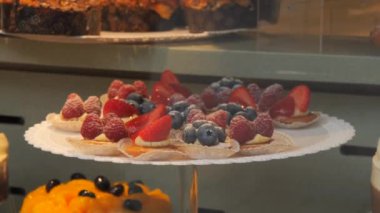Tezgahın üstünde meyve ve böğürtlen olan bir sürü farklı lezzetli pasta. İnsanlar dükkanın camına yansıyor..