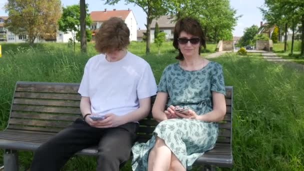 大人の母親とティーンエイジャーの息子が夏の公園のベンチに座ってスマートフォンを見て 画面を押し 携帯電話を手に持って — ストック動画