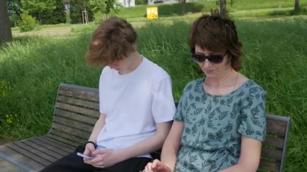 大人の母親とティーンエイジャーの息子が夏の公園のベンチに座ってスマートフォンを見て 画面を押し 携帯電話を手に持って — ストック動画