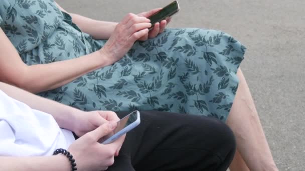 一位成年母亲和一位未成年儿子坐在夏季公园的长椅上 看着智能手机 按下屏幕 手里拿着手机 — 图库视频影像
