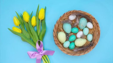 Paskalya Hareketi animasyonu. Mavi tonlarda çeşitli Paskalya yumurtaları hasır sepetin içinde mavi arka planda kuş tüyleriyle narin mor kurdeleli güzel sarı lalelerin yanında hareket eder.