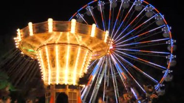 Bir gece eğlence parkında atlıkarınca üzerinde insanların farklı renklerde parlayan dönme dolaba bindiği yavaş çekim..