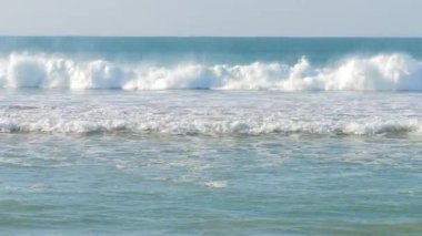 Açık bir gökyüzünün altındaki kumlu bir sahile vuran yumuşak okyanus dalgalarının rahatlatıcı görüntüleri. Turkuaz su güneş ışığında parlayarak huzurlu ve rahatlatıcı bir sahil manzarası yaratıyor. Portekiz 'de çekildi