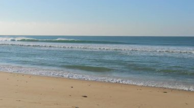 Açık bir gökyüzünün altındaki kumlu bir sahile vuran yumuşak okyanus dalgalarının rahatlatıcı görüntüleri. Turkuaz su güneş ışığında parlayarak huzurlu ve rahatlatıcı bir sahil manzarası yaratıyor. Portekiz 'de çekildi