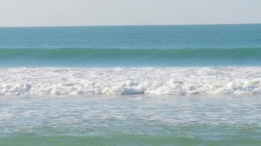 Açık bir gökyüzünün altındaki kumlu bir sahile vuran yumuşak okyanus dalgalarının rahatlatıcı görüntüleri. Turkuaz su güneş ışığında parlayarak huzurlu ve rahatlatıcı bir sahil manzarası yaratıyor. Portekiz 'de çekildi