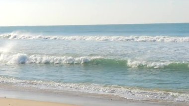 Açık bir gökyüzünün altındaki kumlu bir sahile vuran yumuşak okyanus dalgalarının rahatlatıcı görüntüleri. Turkuaz su güneş ışığında parlayarak huzurlu ve rahatlatıcı bir sahil manzarası yaratıyor. Portekiz 'de çekildi