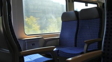 Mavi desenli koltukları ve tahta kollukları olan boş bir tren vagonunun huzurlu bir yavaş çekim görüntüsü. Geniş pencerelerden manzaralı bir kırsal alan yavaşça geçiyor ve rahatlatıcı bir manzara yaratıyor.