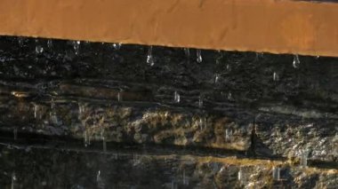 Detaylı bir yakın plan su, bir taş çeşmenin kenarından yavaşça akarak, aşağıdaki dokulu yüzeye dökülen hassas damlacıklar oluşturur. Ağır çekimde yakalanan, hareketin