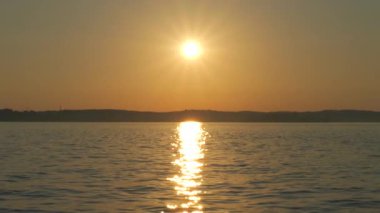 Huzurlu bir gölün üzerinde büyüleyici bir günbatımı, altın güneş hafif dalgalı su üzerinde parıldayan bir yansıma oluşturur. Sıcak akşam ışığı huzurlu ve huzurlu bir atmosfer yaratır.