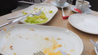 Açık bir kafedeki ahşap bir masanın üzerindeki boş ve kısmen boş tabakların yakın plan görüntüsü, arta kalan salata, kırıntılar ve çatal bıçaklarla birlikte. Gövdede iki fincan çay tabağı da görülebilir.