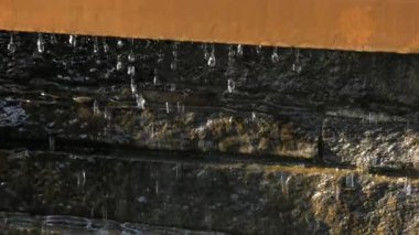 Detaylı bir yakın plan su, bir taş çeşmenin kenarından yavaşça akarak, aşağıdaki dokulu yüzeye dökülen hassas damlacıklar oluşturur. Ağır çekimde yakalanan, hareketin