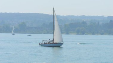 Tamamen geniş yelkenli güzel beyaz bir yelkenli sakin mavi göl sularında zarif bir şekilde süzülüyor. Arka plandaki yumuşak sis, bereketli manzaraya derinlik katıyor.