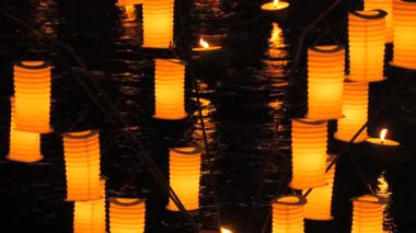 Narin dallardan sarkan parlak kırmızı kağıt fenerlerin olduğu çarpıcı bir gece sahnesi, nazikçe akan bir su yüzeyinde güzelce aydınlatılmış. Yüzen mumlar ılık ve büyüleyici bir parıltı ekler, dalgalanan suya hafifçe yansıtır.