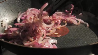 Kızartma tavasında cızırdayan kırmızı soğan aşırı yakın plan, bir spatulayla hafifçe karıştırılmış. Sahne yavaş çekimde çekiliyor, pişirme işlemi sırasında dokuları, buharı ve parlayan yağı vurguluyor. Yemek içeriği ve yemek tarifi için ideal