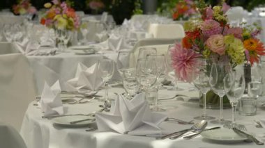 Düğün resepsiyonunda çok güzel dekore edilmiş bir masa. Beyaz çarşaflar, düzgünce katlanmış peçeteler, şarap bardakları ve çiçekli süsler zarif ve şenlikli bir atmosfer yaratır.