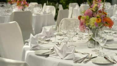 Resmi bir etkinlik için masanın ortasına yerleştirilmiş parlak ve renkli çiçek aranjmanı. Beyaz çarşaflar, cam eşyalar ve zarif sofra takımlarıyla çevrili.