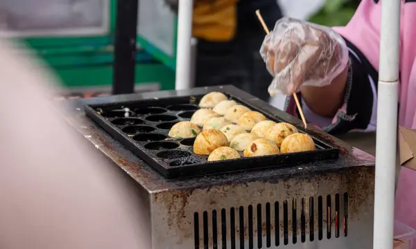A man is cooking takoyaki. Traditional food on a grill with a bottle of ketchup authentic from japan. The food is small and round, and the man is wearing gloves