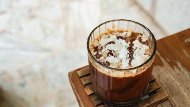 Hindistan cevizli buzlu kahve uzun bardakta, hindistan cevizi dökülmüş ve kahve çekirdekleri ahşap masada..