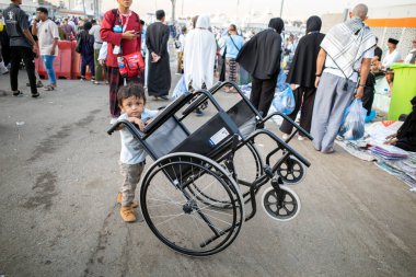 Mekke, Suudi Arabistan - 17 Haziran 2024: Hac sezonunda Suudi Arabistan 'ın Mina kentindeki hediyelik eşya satıcılarının yanında tekerlekli sandalyeyle oynayan küçük bir çocuk.