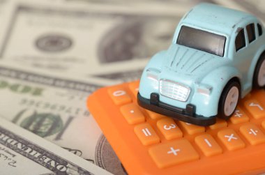 Turuncu hesap makinesinin üzerinde duran mavi bir oyuncak araba, Amerikan dolarlarıyla çevrili. Sigorta, bakım ve akaryakıt maliyetleri gibi araba mülkiyetine ilişkin mali hususlar.