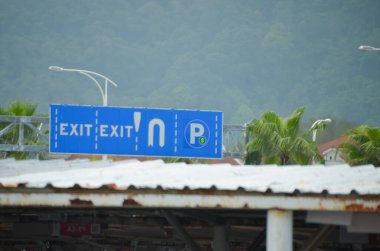 Açık mavi arka planı ve beyaz harfleriyle tepedeki tabela sürücülere çıkışa ve belirlenmiş park alanına doğru rehberlik ediyor..