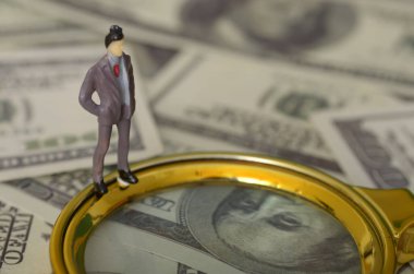 Minyatür bir işadamı büyüteçle bir yığın banknotu inceliyor. İş adamı belli bir şey arıyor gibi görünüyor, belki bir fırsat ya da bir meydan okuma..