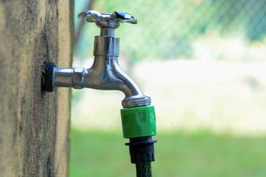 Görüntü, üzerine yeşil bir hortum bağlanmış bir duvara bağlı metal bir musluğun yakın çekimini gösteriyor..