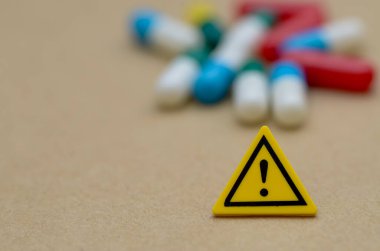 Renkli hapların bulanık arka planına karşı sarı uyarı işareti net bir şekilde odaklanmıştır. Yan etkilerin, aşırı dozun ve reçeteli istismarın risklerini sembolize etmektedir..