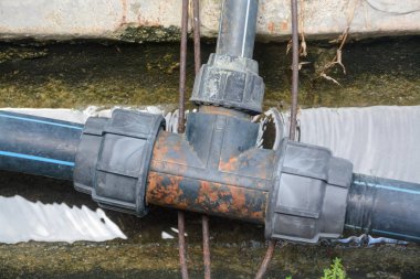 Rutubetli beton bir kanalda paslı, yıpranmış siyah plastik bir T-kavşağı acil tesisat bakımını sembolize eder ve su sızıntısı riskini vurgular..