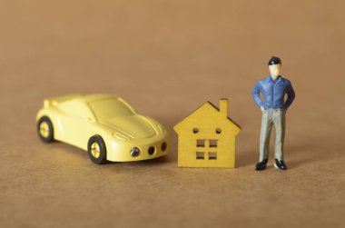A figurine man stands proudly next to a wooden house and a sports car, symbolizing personal assets, wealth, and major life goals like home and car ownership.