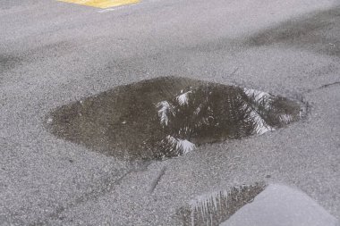 Yağmur suyuyla dolu tehlikeli bir çukur, kötü yol koşullarını ve tropikal iklimde acil altyapı bakım ihtiyacını vurgulayan bir palmiye ağacını yansıtır..