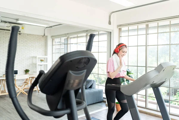 Cute Asian woman runs on a treadmill in the gym and wears red headphone and wave hand for greeting someone, the girl listens to music during a cardio workout for weight loss, Sport exercise and music concept.