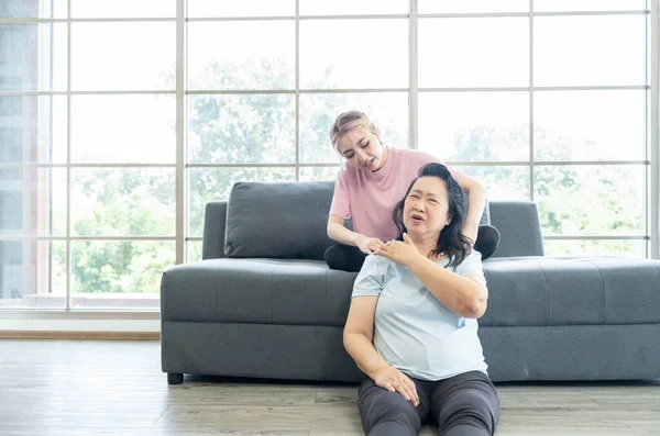 Asian mother fill pain and young Asian daughter sit on sofa and giving massages shoulder, relieves pain of elderly Asian mother at home.