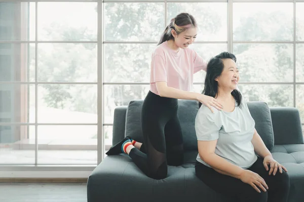 Asian mother fill happy with young Asian daughter sit on sofa and giving massages her shoulder, relieves pain of elderly Asian mother at home.