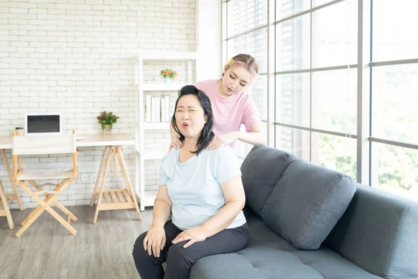 Asian mother fill good at face that young Asian daughter sit on sofa and giving massages her shoulder, relieves pain of elderly Asian mother at home.