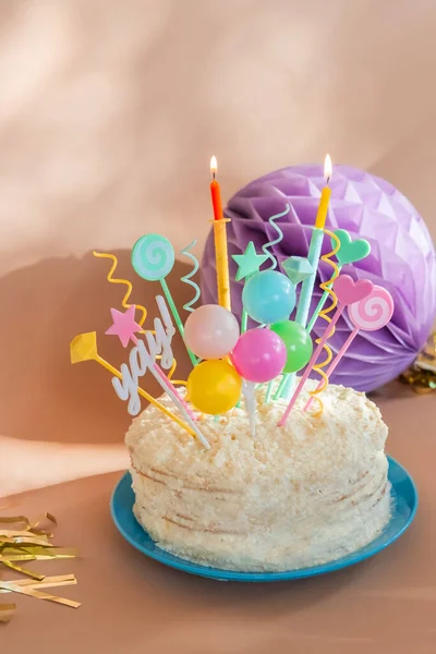 Cream birthday cake with candles, paper balloons, fringe and confetti. Party decorations and treat.