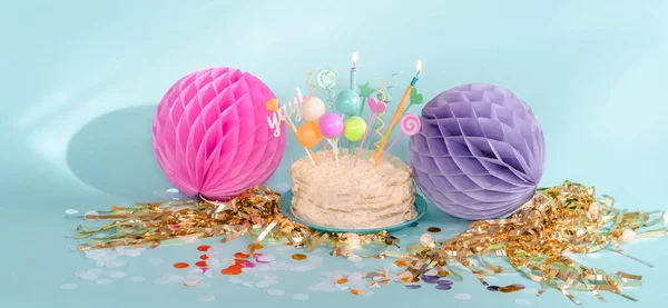 Cream cake with candles. Party decor with fringe, paper balloons, poms and confetti.
