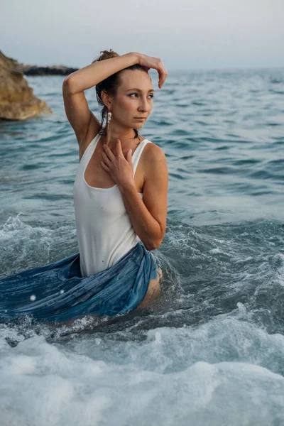 Beautiful young woman in top and skirt posing in the sea waves in the evening. Fashion photo shoot in nature.