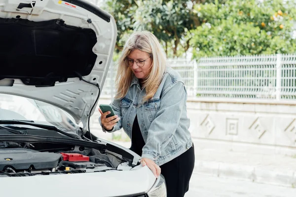 Woman near car with an open hood making video call to car service from mobile phone.Traveler girl, faced with a broken rental car, asking for help with a mobile phone.
