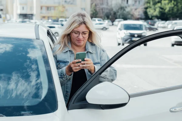 Woman standing near car and looking at her smartphone, paying for parking and navigates the city.