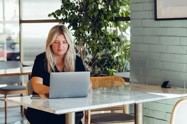 stock image Middle-aged blond woman working with laptop while sitting in cafe indoors.