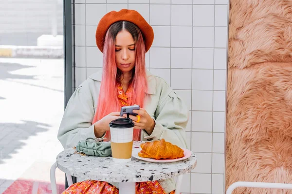 Young woman with unique fashion style having breakfast in cafe and using mobile phone.