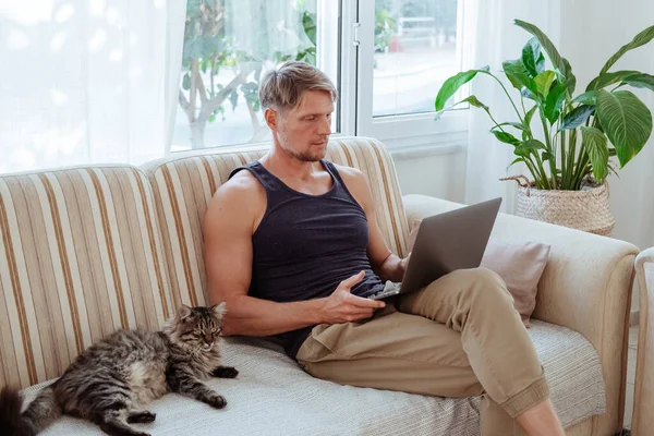 Man sits with a laptop near cat on the couch at home. Free time with technology. High quality photo