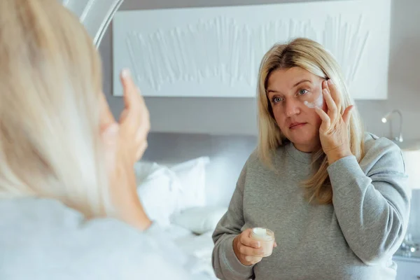Attractive young plus size blonde woman looking at mirror touching her face. Middle-aged woman doing make -up, enjoy healthy skin care, aging beauty, skin treatment cosmetics concept.