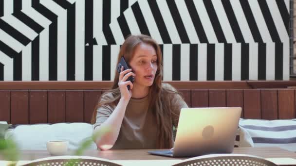 若い女性が携帯電話で話をし コワーキングスペースでコンピュータ上で動作します フリーランスの女の子は電話で話すと現代のカフェでラップトップ上のソーシャルメディアをチェック — ストック動画