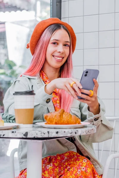 Young woman with unique style having breakfast in cafe and using mobile phone. Teenagers girl with colorful hair chatting on cellphone.