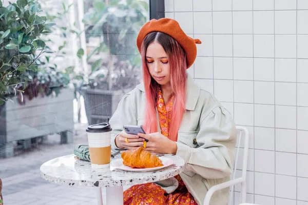 Young woman with unique style having breakfast in cafe and using mobile phone. Teenagers girl with colorful hair chatting on cellphone.