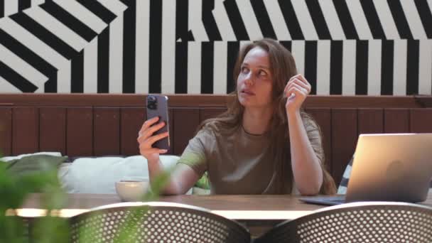 若い女性はコワーキングスペースで携帯電話について話します フリーランスの女の子持っていますビデオチャットと電話で現代的なカフェ — ストック動画