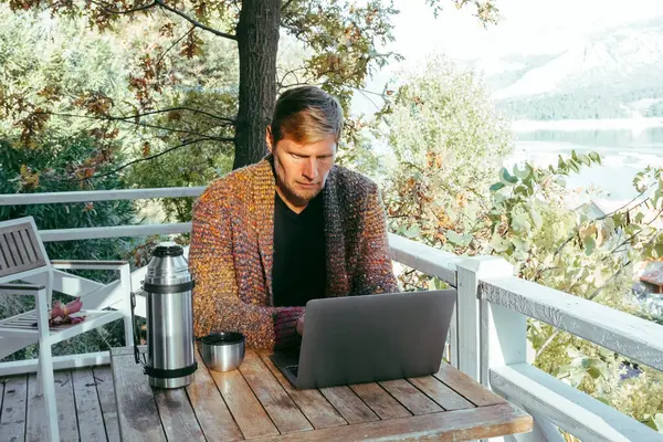 Mature man in knitted cardigan drinking coffee on cozy wooden balcony and tipping on laptop computer. Autumn weekend vibe.